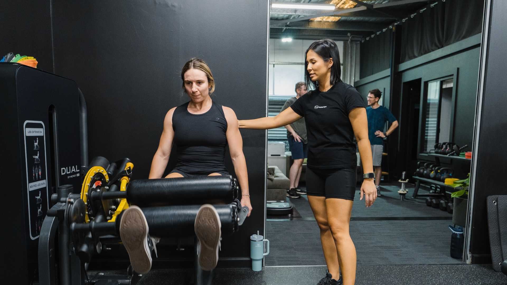 Trainer guiding a woman on a seated leg extension machine in a gym, with other people and equipment visible in the background.