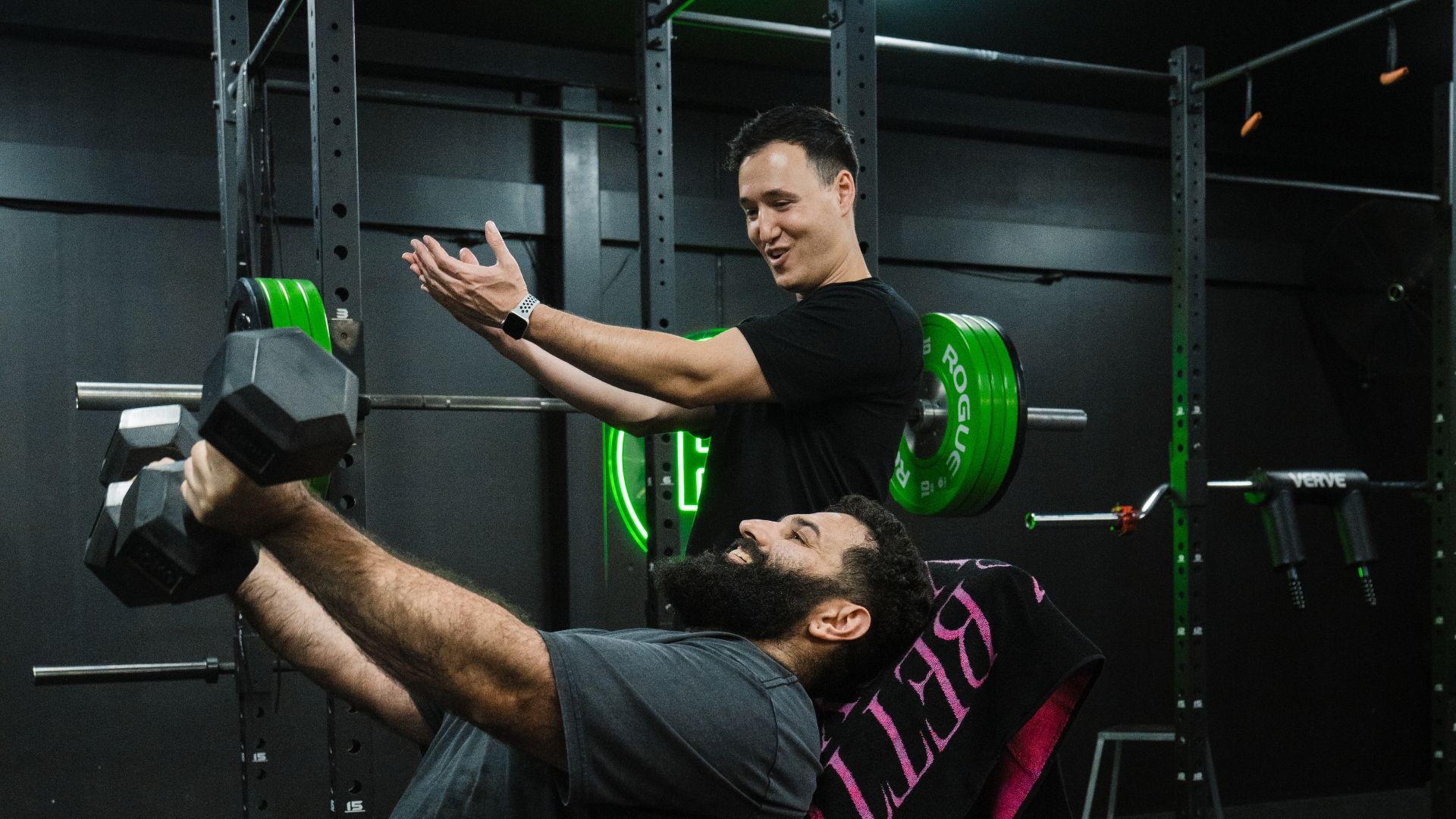 Trainer coaching a man performing an incline dumbbell press in the gym, both smiling as he lifts the weights.
