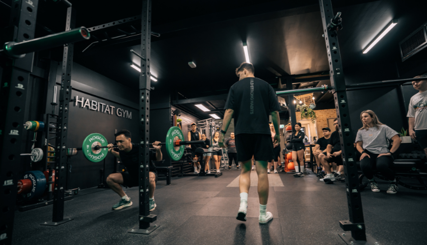 Man doing barbell squats at Habitat Gym’s weight area while being guided by a personal trainer