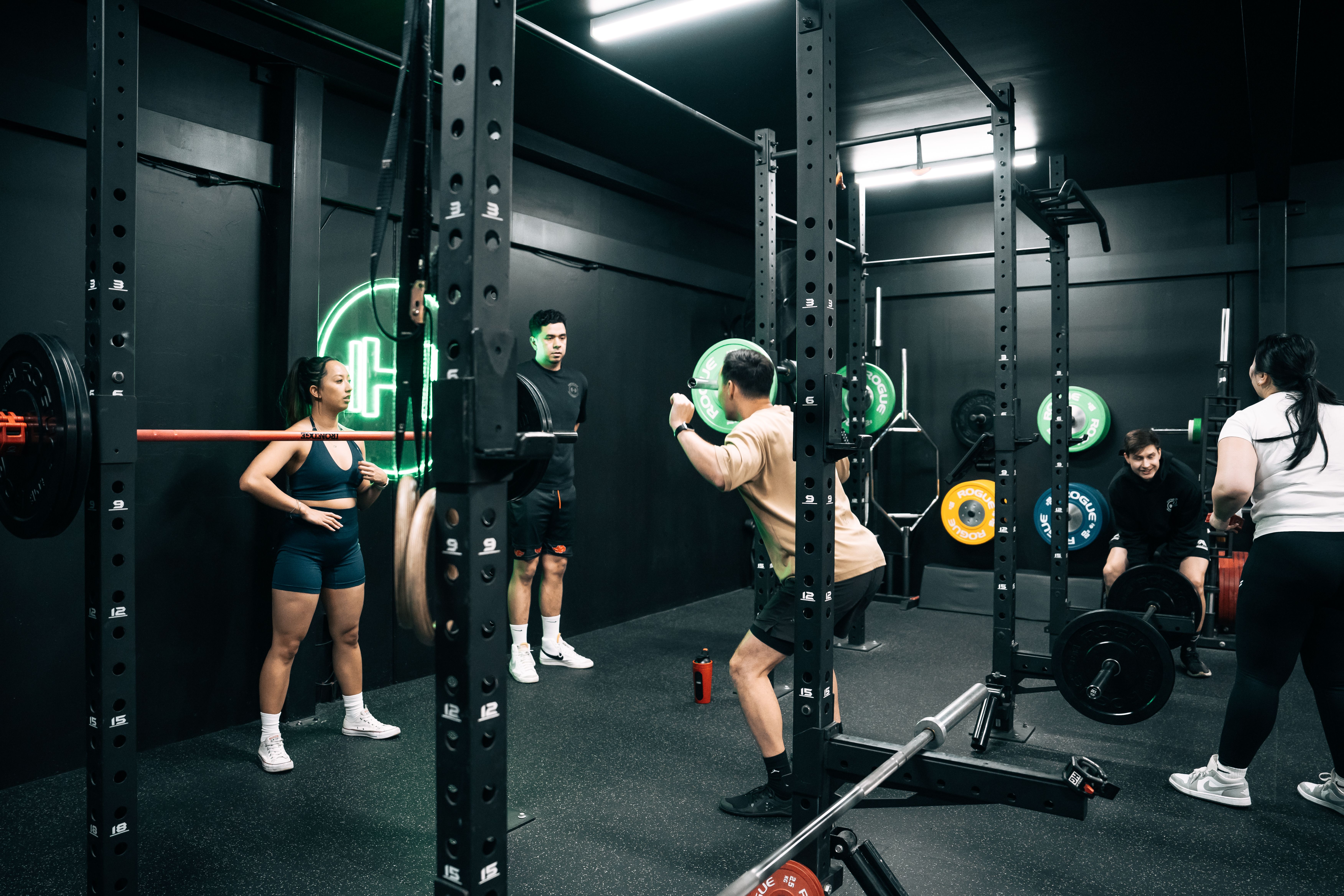 A group of people lifting weights at Habitat Gym. One man is performing a barbell back squat inside a squat rack, while others observe or train nearby. The gym has a black interior with neon lighting and coloured Rogue weight plates. A female coach stands to the left, watching attentively.