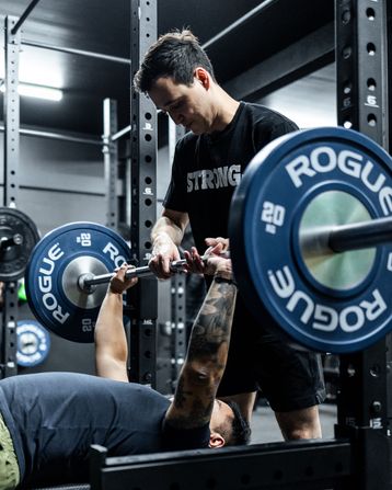 Trainer spotting client on bench press at Habitat Gym using Rogue Fitness equipment