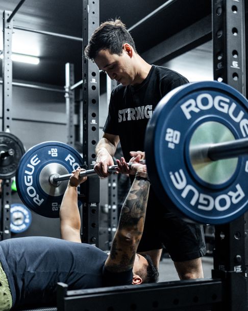 Trainer spotting client on bench press at Habitat Gym using Rogue Fitness equipment