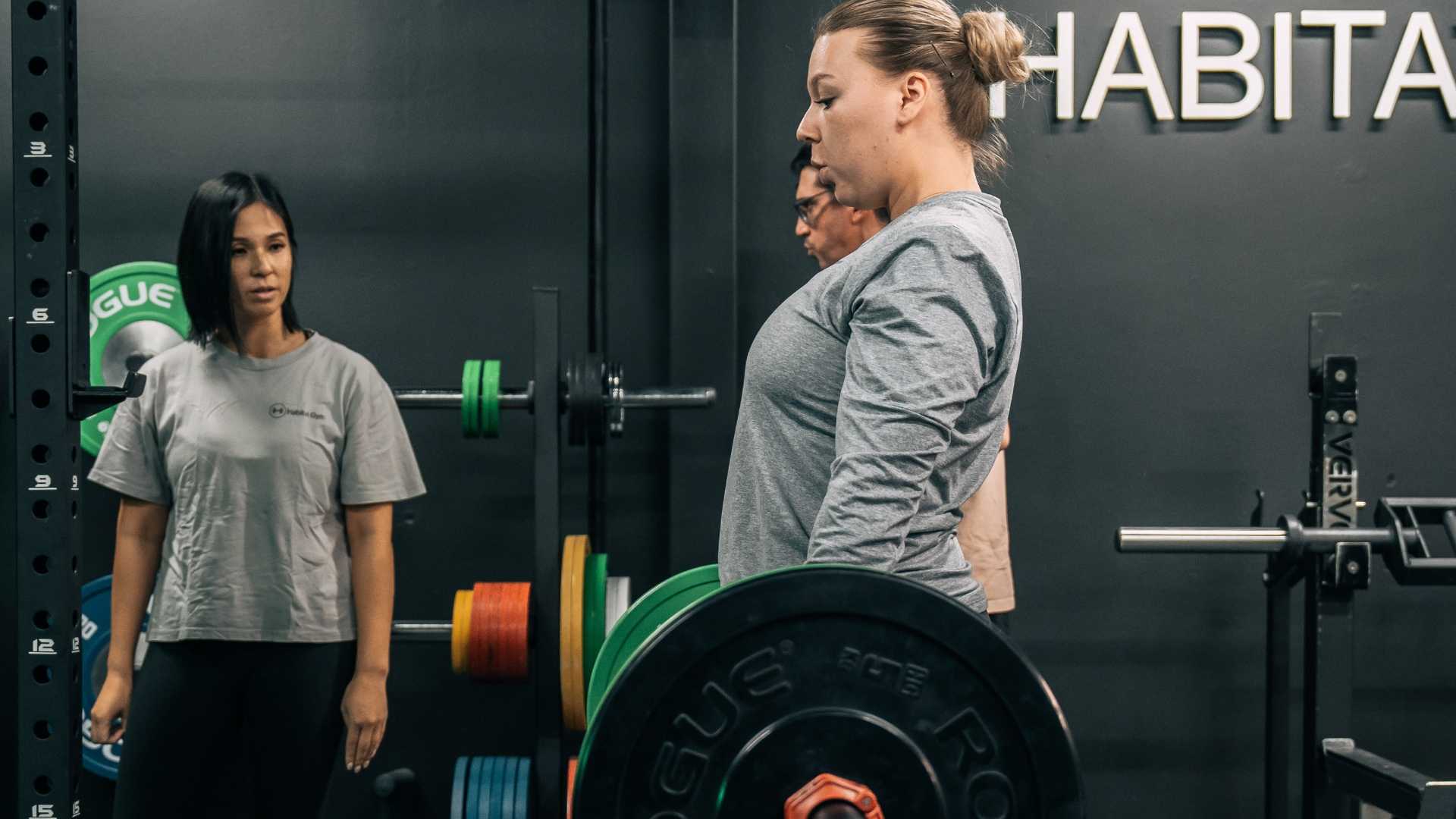Female client performing a deadlift with a barbell under the supervision of a personal trainer in a gym setting.