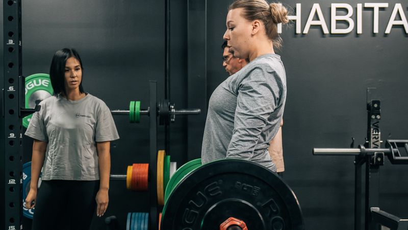 Female client performing a deadlift with a barbell under the supervision of a personal trainer in a gym setting.