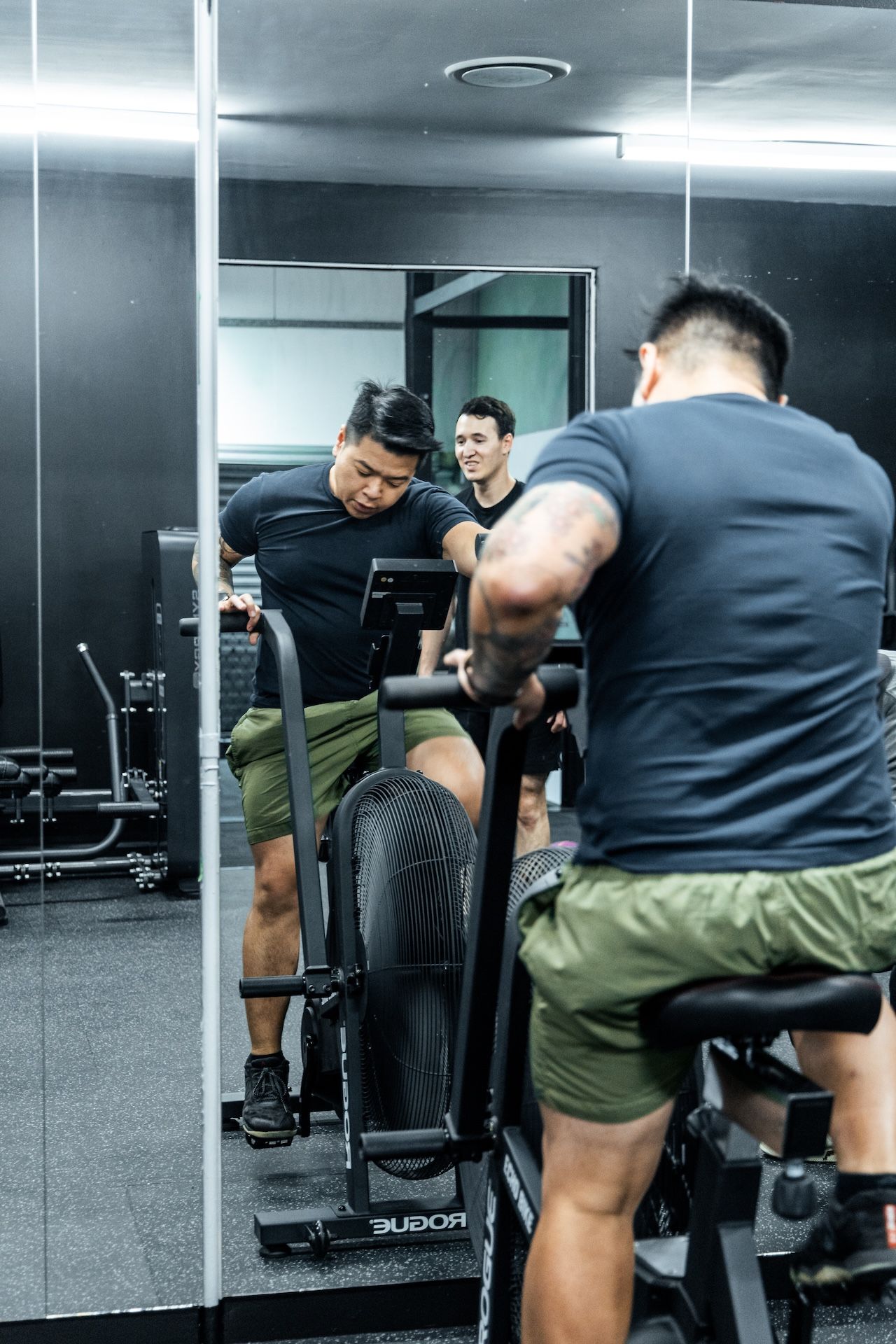 Male gym member riding Rogue Echo bike during intense cardio interval with trainer guidance