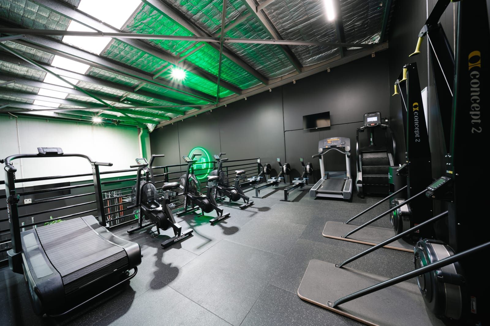 Mezzanine cardio floor under green-lit rafters with a curved treadmill, Rogue Echo bikes and Concept2 ergs