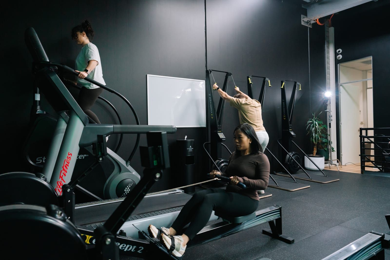 Member mid-pull on a wall-mounted SkiErg while others use the Concept2 rower and stair climber