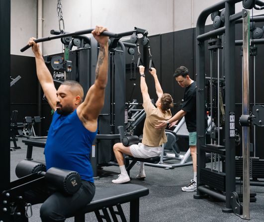 Members training on the pin-loaded machines while a coach fine-tunes one member’s lat pulldown form at Habitat Gym