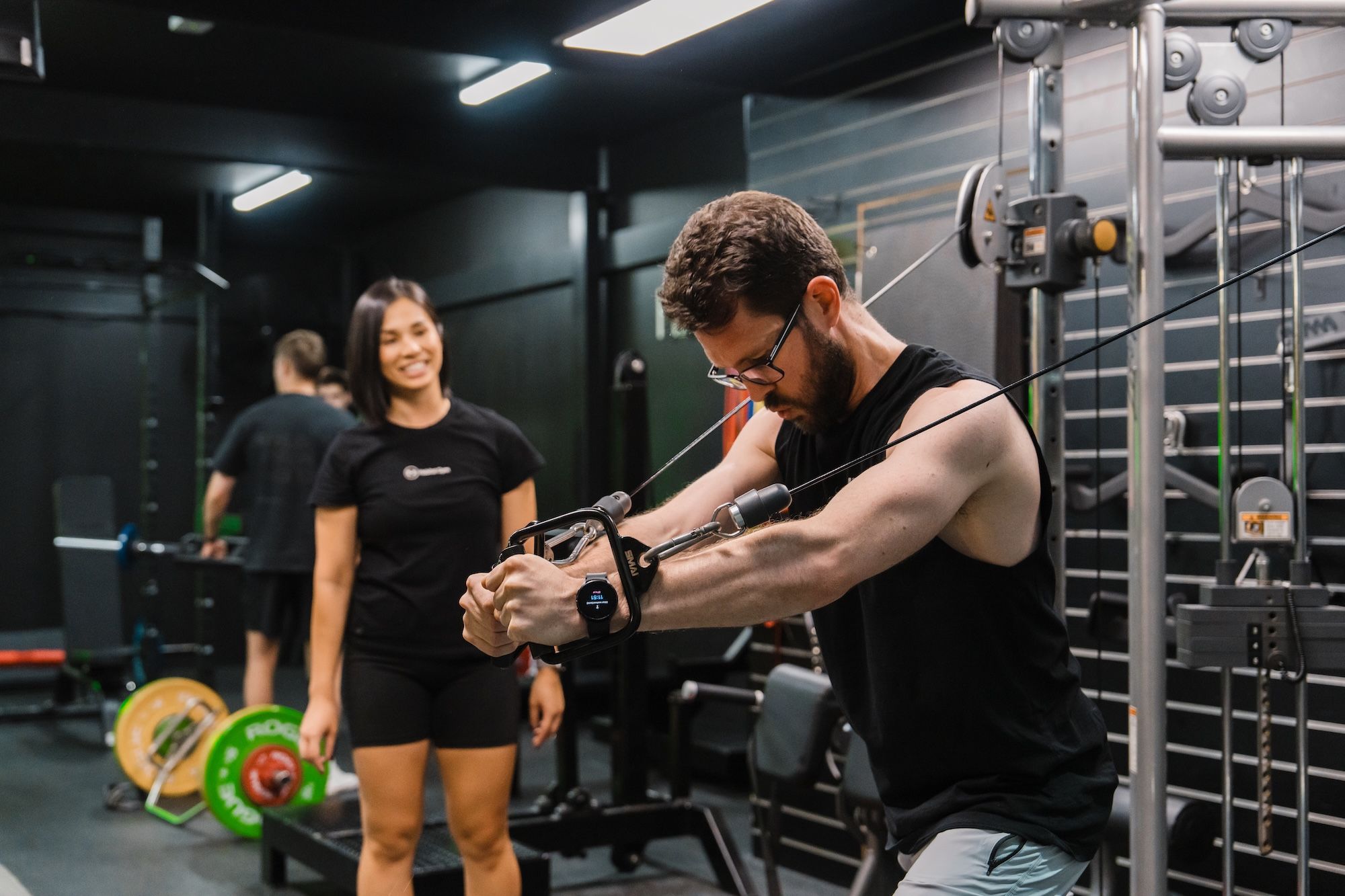 Man in a black tank top and glasses performing a standing cable chest fly on a dual cable machine, while a coach in a black t-shirt stands behind him smiling. Other members train in the background of the gym floor.