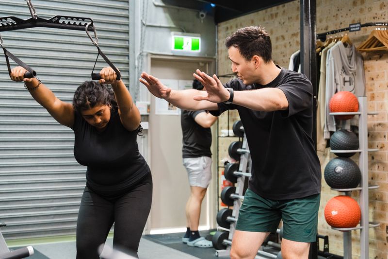 Coach guiding a client through a cable exercise with hands-on form correction during a semi-private personal training session in a gym.