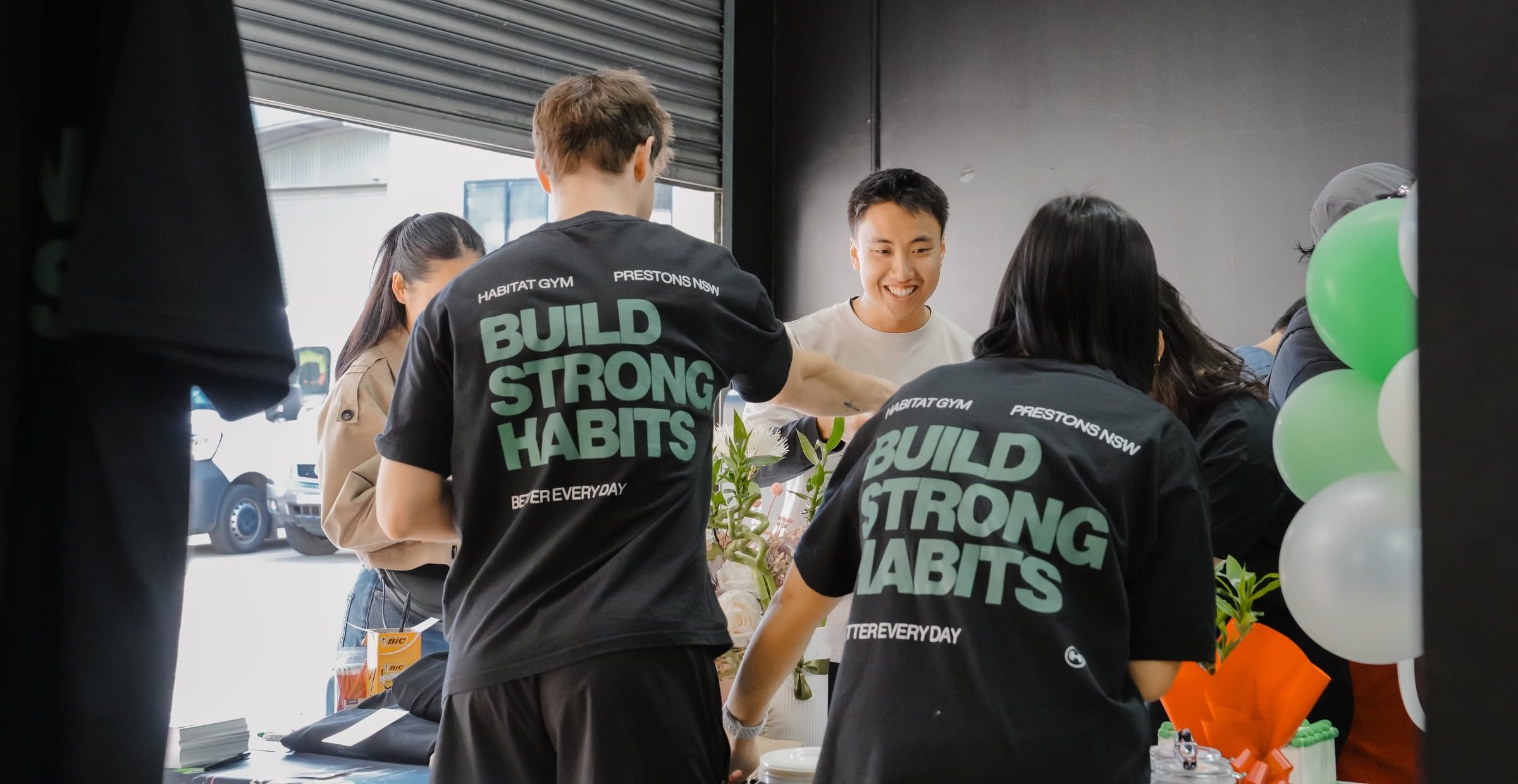Habitat team members in Build Strong Habits shirts welcoming guests at the gym’s open day, balloons strung by the roller door