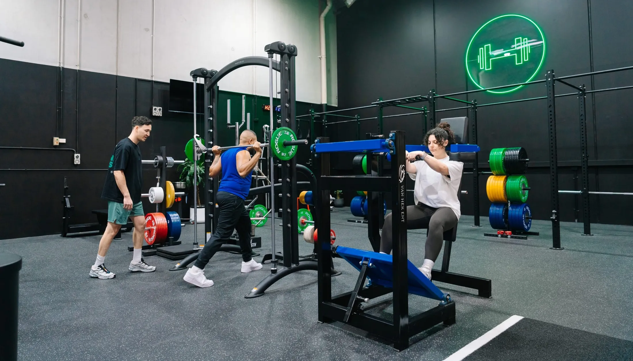 Coach watching a member squat at the Smith machine while another member trains on a plate-loaded machine beneath the green neon dumbbell sign at Habitat Gym in Prestons