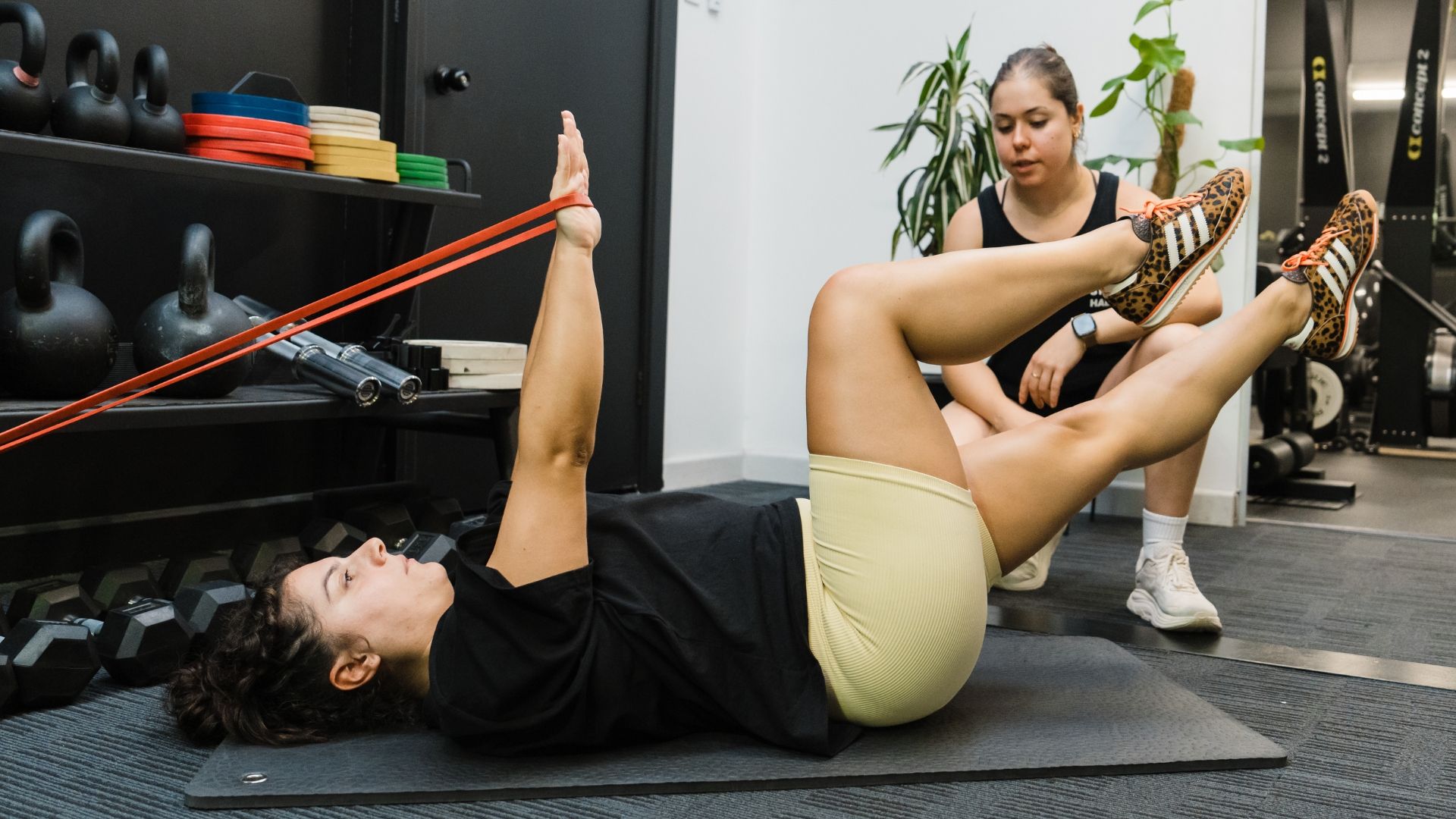 Coach guiding client performing resistance band core exercise on gym floor with kettlebells and equipment in the background
