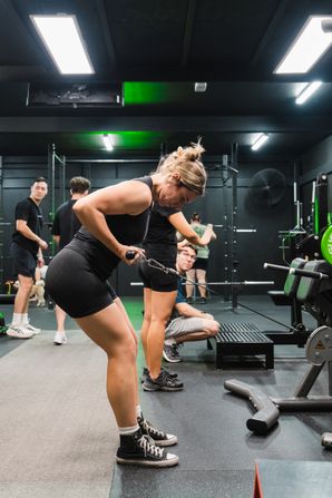 A focused woman performing cable rows at Habitat Gym, hinged forward with her elbow drawn back. Other members train in the background under bright overhead lights.