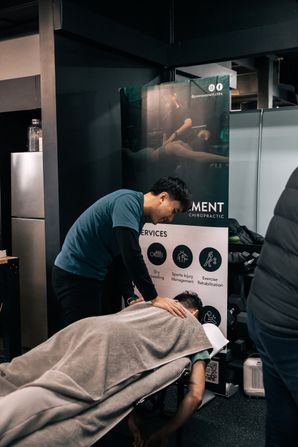 Chiropractor in a blue t-shirt working on a client lying face-down on a treatment table inside the gym, with a Pro Movement Chiropractic banner behind them listing services like dry needling, sports injury management, and exercise rehabilitation.