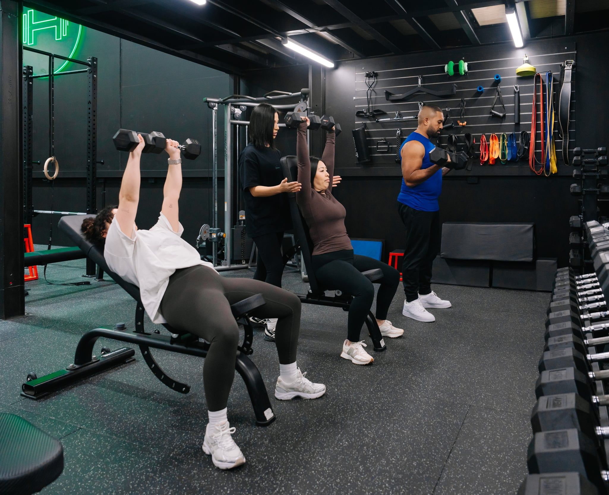 Personal trainer spotting a client’s seated dumbbell shoulder press while other members train beside the dumbbell rack at Habitat’s private gym in Prestons, Sydney