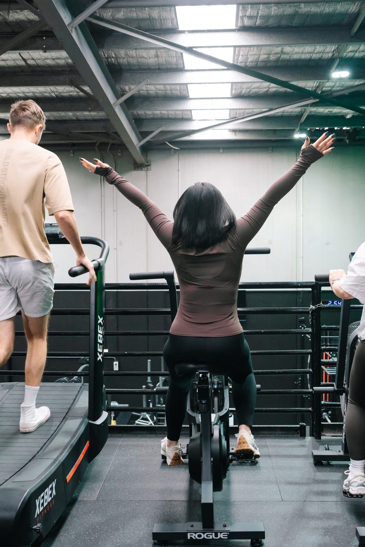 Members training side by side on a curved treadmill and a Rogue Echo bike, one stretching her arms overhead