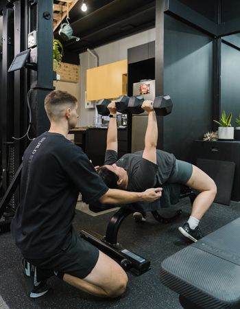 Personal trainer supporting a gym member whilst doing arm presses with free-weights inside Habitat Gym