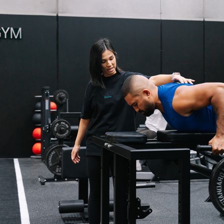 Coach guiding a member into position on a bench, with medicine balls and weight plates racked behind