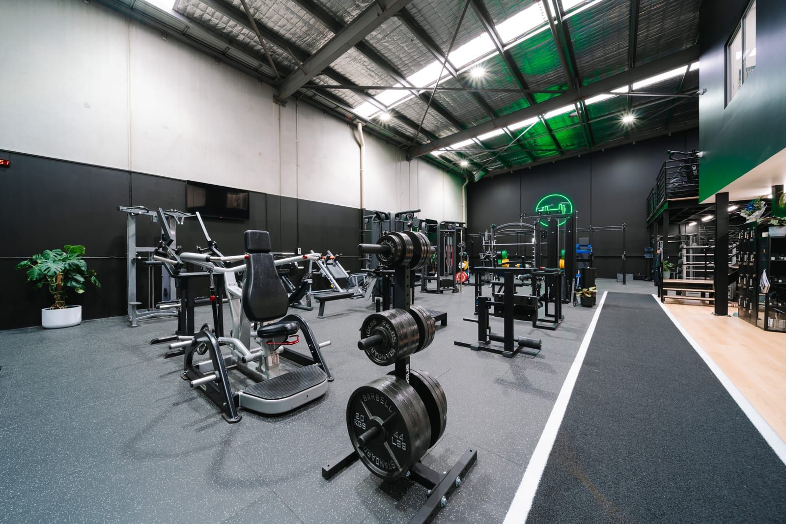 Wide view of the strength zone with plate-loaded machines, weight trees and the green neon dumbbell sign on the far wall