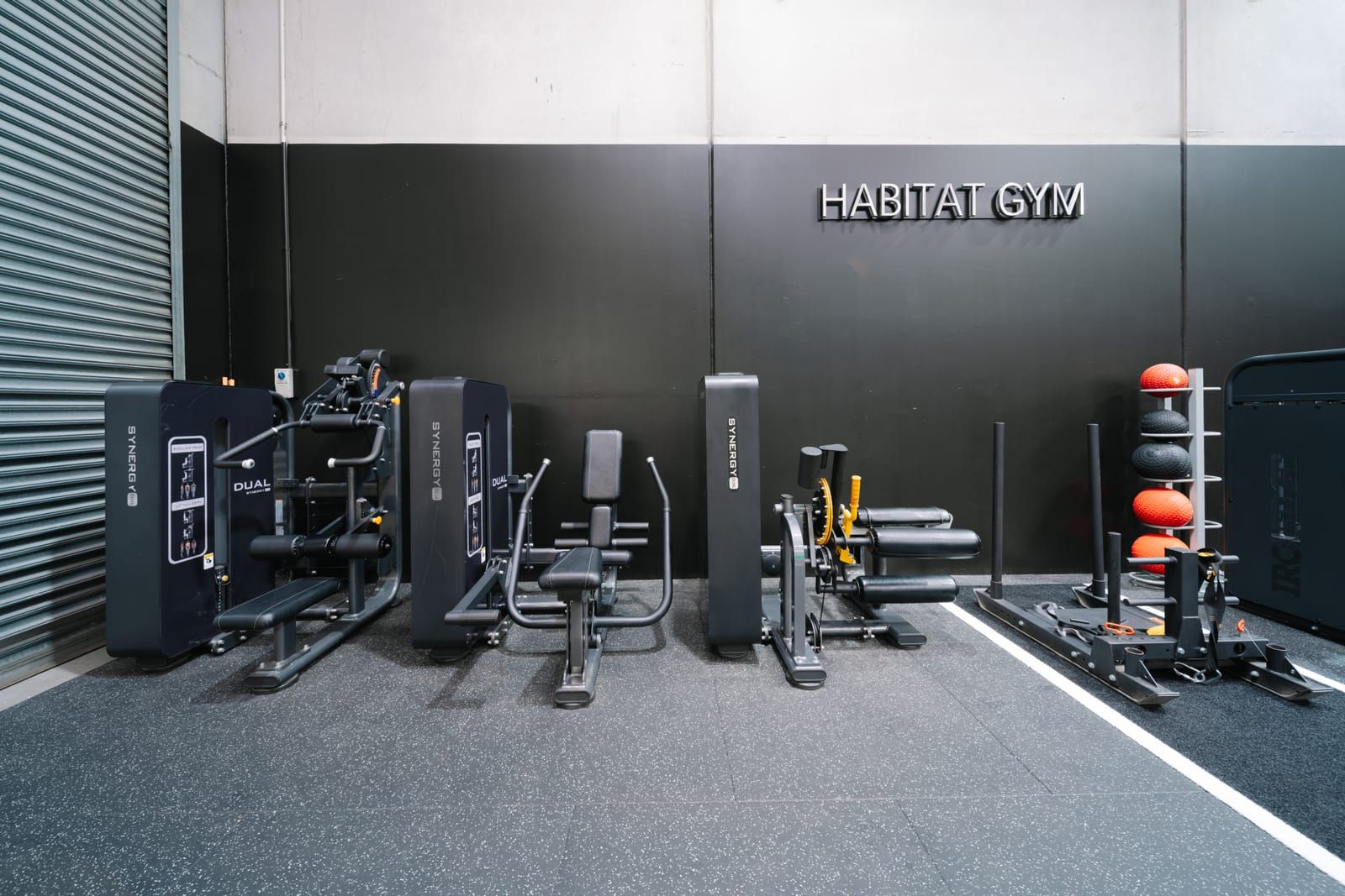 Line of pin-loaded leg machines beneath Habitat Gym wall lettering, with medicine balls racked alongside