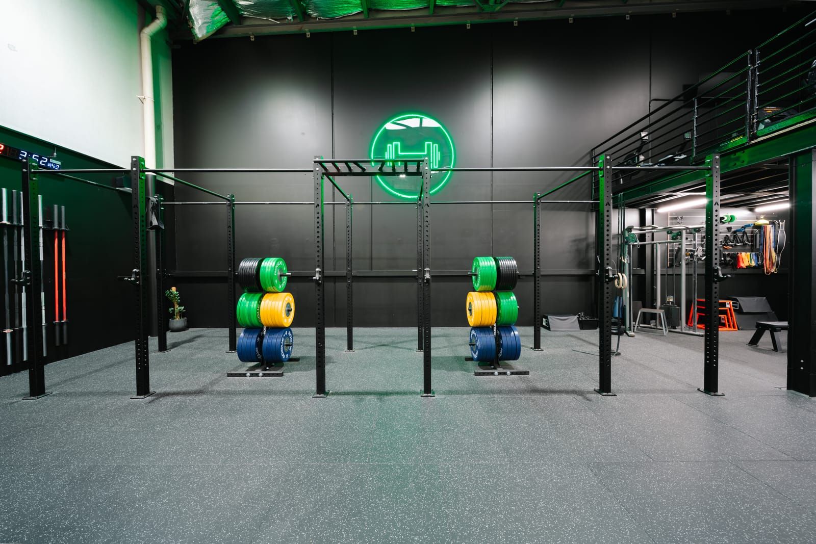 Free-standing rig with pull-up bars and stacked bumper plates beneath the green neon dumbbell emblem