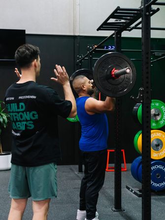 Coach cueing a member holding a loaded barbell in the front rack position at the rig, Rogue bumper plates stored nearby