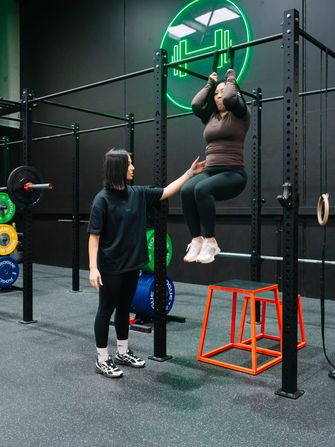 Coach steadying a member through hanging knee raises on the rig beneath the green neon dumbbell sign at Habitat Gym