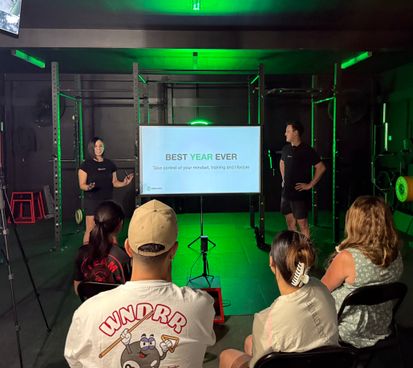 Two Habitat Gym coaches presenting a 'Best Year Ever' workshop to a small group of seated members inside the gym, with a projected slide reading 'Take control of your mindset, training and lifestyle.'