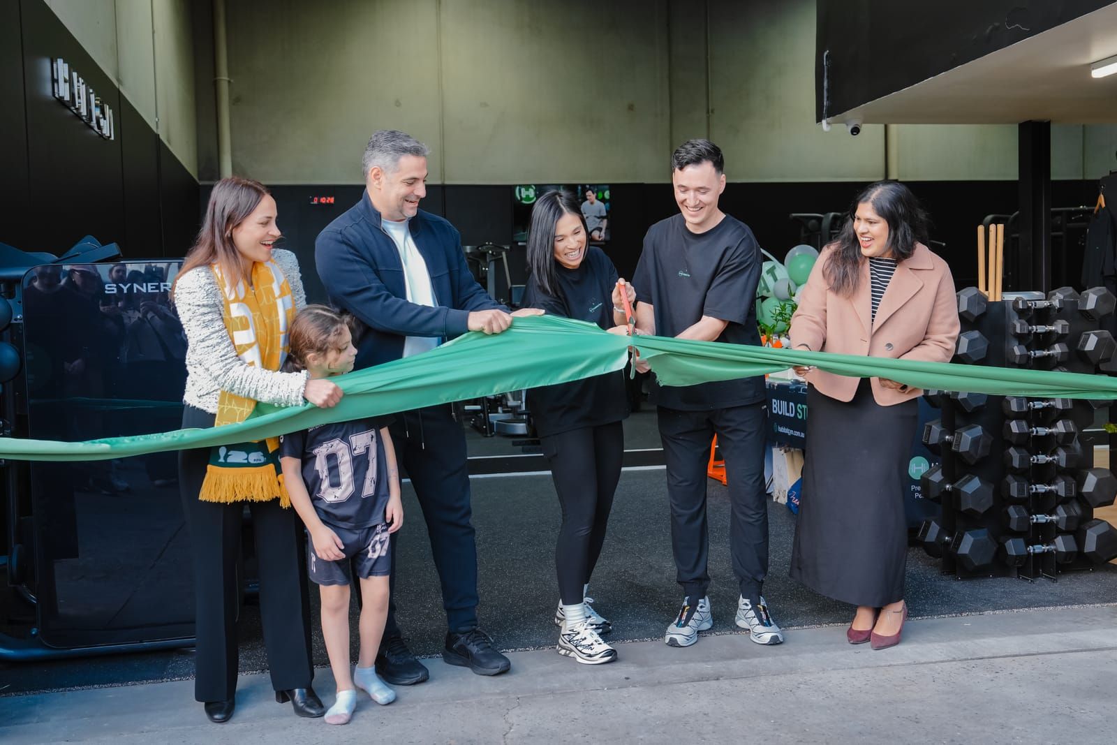 Guests cutting a wide green ribbon in front of the dumbbell racks at Habitat Gym’s grand opening
