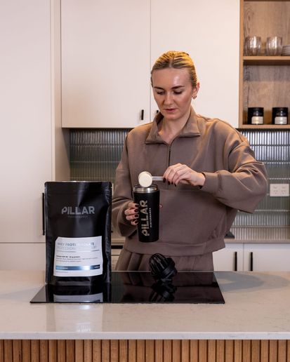 Woman preparing a Pillar Performance recovery supplement in a kitchen, with a shaker and pouch on the bench